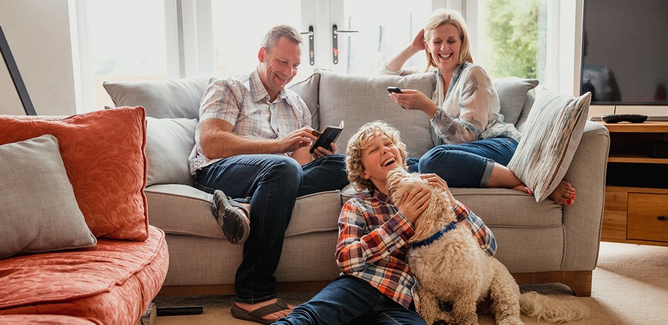 A happy family is sitting in the living room. Their son is sitting on the floor with the family dog licking his face.