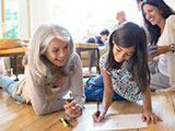 A grandmother and granddaughter coloring on the floor of their living room.