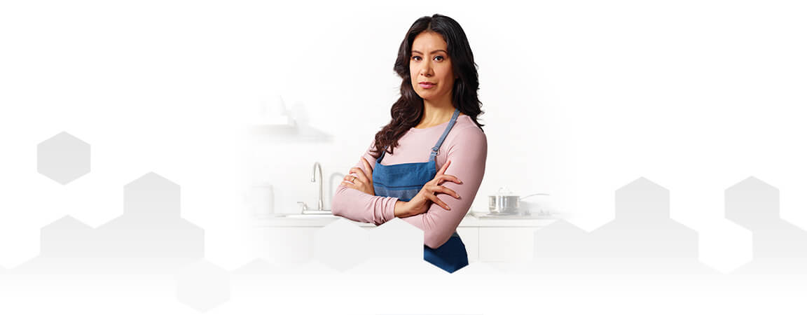 Una mamá con delantal parada junto a la encimera de la cocina con los brazos cruzados.